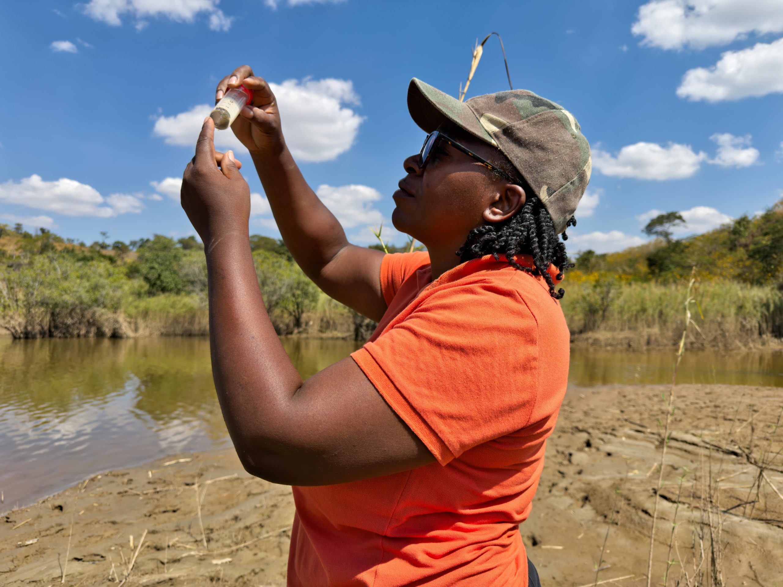 National Geographic Explorer Dr. Moola Mutondo at a river in Zambia’s Copperbelt, examining a vial full of green photosynthetic microbes she just collected from the river